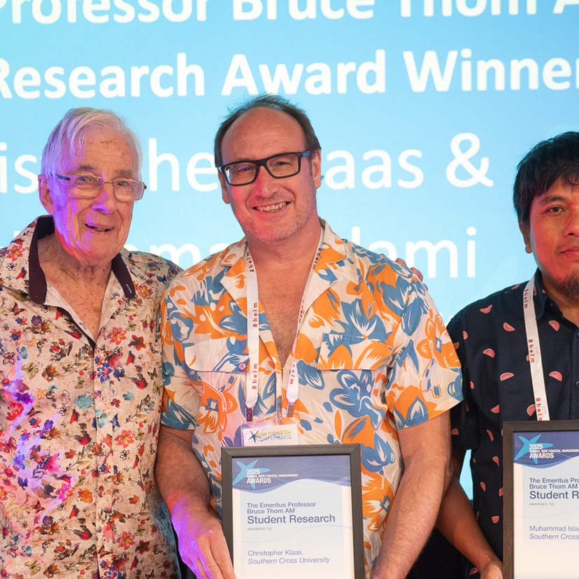 3 men on a stage in front of a screen, 2 of them holding awards