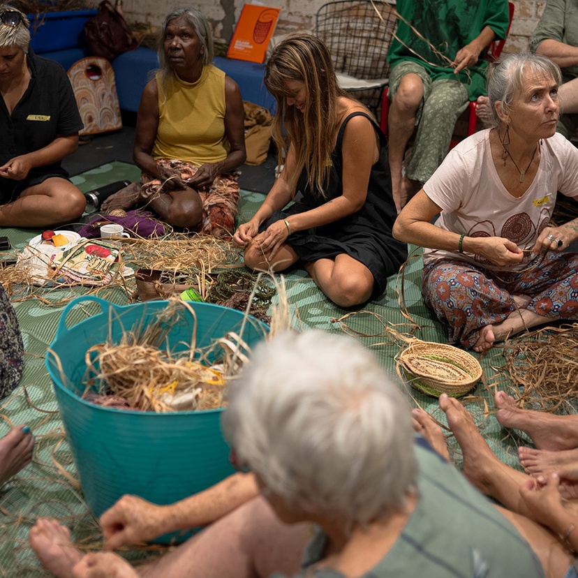 A group of Indigenous and non-Indigenous women sitting on the floor making string from plant fibres.