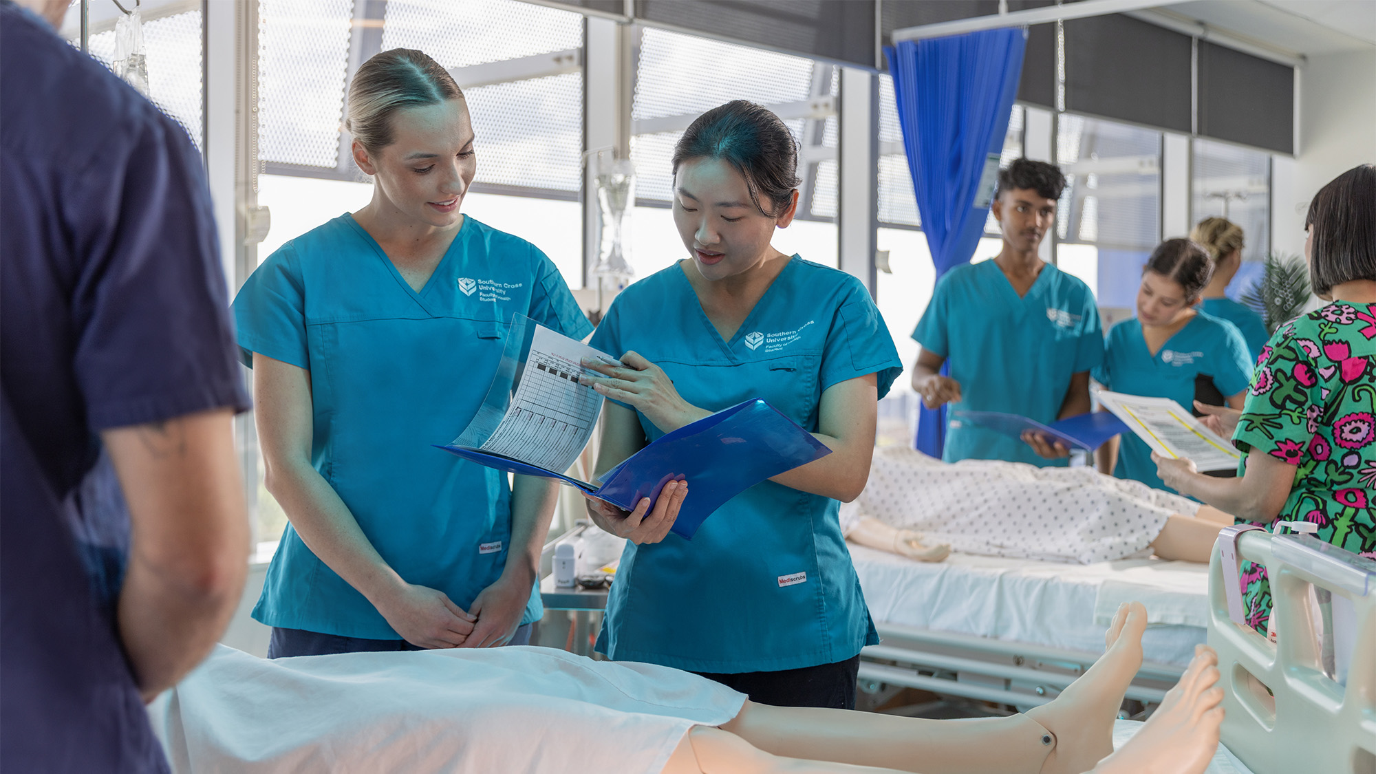 Two students in nursing lab with sim doll on hospital bed