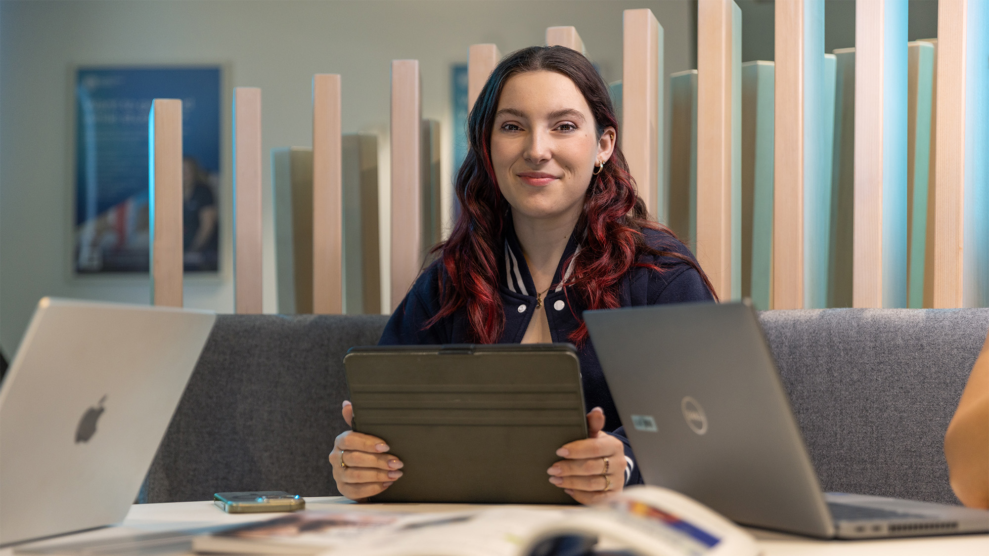 Student with iPad in library and laptops on her left and right