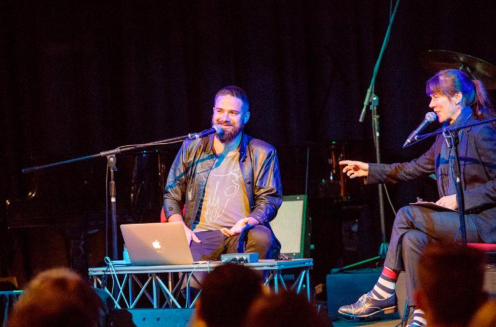 Events and News A man with a laptop and musical instruments talking to a woman on stage