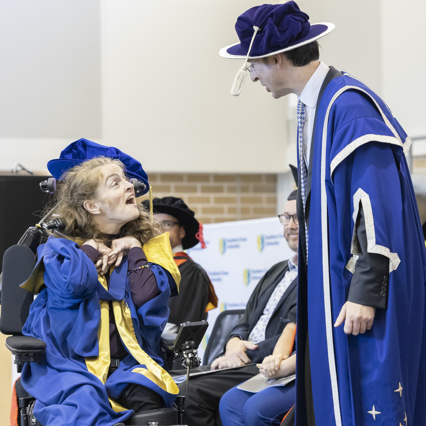 A woman wearing academic gown in a wheelchair exchanging conversation with a man also in academic gown.