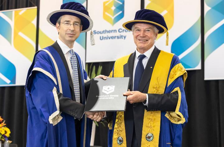 Two men in academic gowns shaking hands in front of Southern Cross University banners