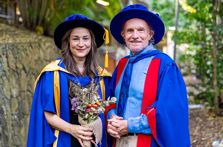 A woman and man both in graduation gowns, with the woman holding a bouquet of flowers