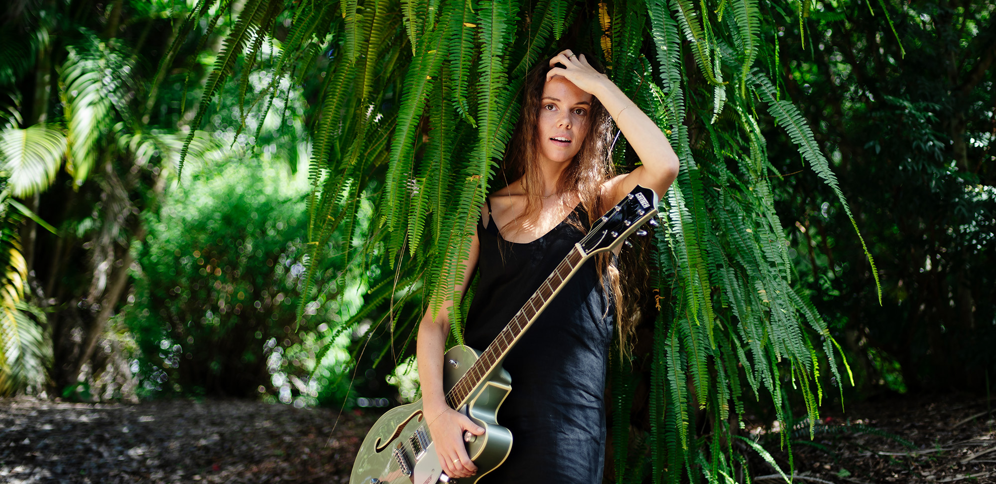 A student stands holding a guitar and  is pushing her long hair back. There is a backdrop of ferns, at the Southern Cross Univerist Lismore Campus